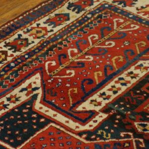 Geometric red and dark blue patterned rug with cream borders rests on light brown hardwood flooring.