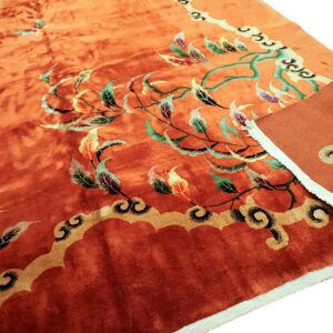 Deep orange rug featuring green, yellow, and maroon botanical patterns and a scalloped border, with the edge folded.