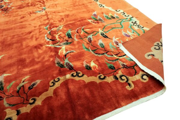 Deep orange rug featuring green, yellow, and maroon botanical patterns and a scalloped border, with the edge folded.