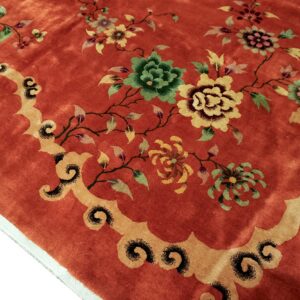 Reddish-orange rug featuring green and yellow floral designs bordered by tan scrollwork and visible pile texture.