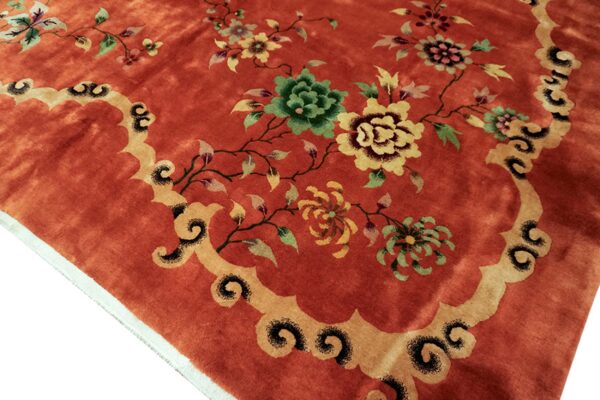 Reddish-orange rug featuring green and yellow floral designs bordered by tan scrollwork and visible pile texture.