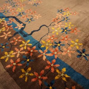 Brown rug with a stylized tree branch, blue stream, and colorful scattered yellow, coral, and blue blossoms.