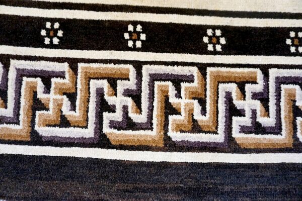 Close-up of a rug border showing interlocking geometric designs and small flowers in cream, tan, and dark colors.