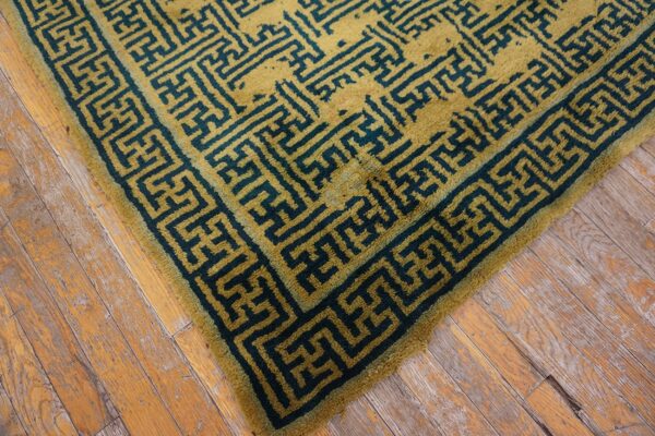 A textured olive green rug with a deep teal geometric fretwork pattern rests on distressed wood floors.