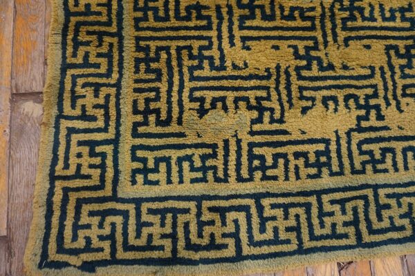 Thick-pile rug corner showing a dark geometric key pattern on an olive gold ground over wood planks.