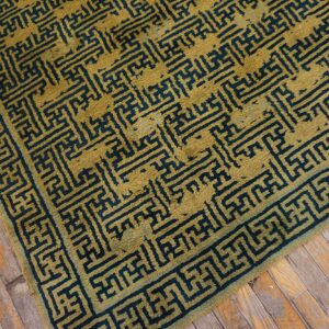 Mustard gold rug with an angular dark teal pattern and border lies on distressed wooden planks.