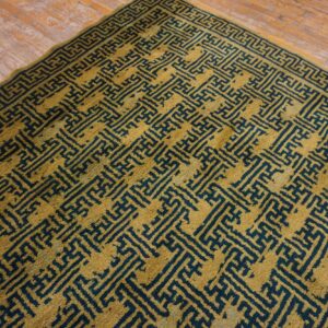 Gold rug with a dense, dark teal geometric key pattern sits on distressed hardwood flooring.