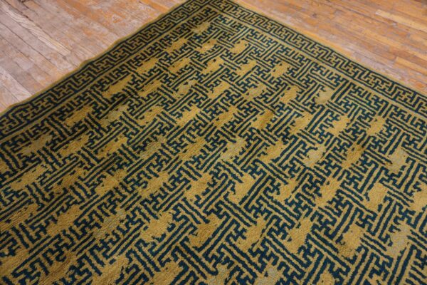 Gold rug with a dense, dark teal geometric key pattern sits on distressed hardwood flooring.