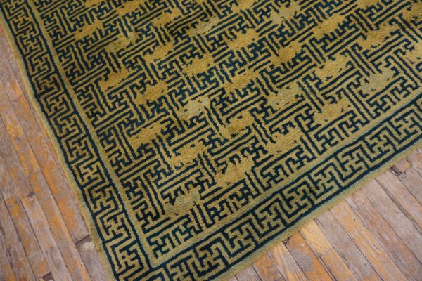 Textured gold rug with an interlocking dark blue maze pattern rests on worn hardwood flooring.