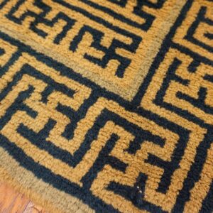 Golden yellow, high-pile rug with a dark blue interlocking key pattern rests on distressed wood planks.