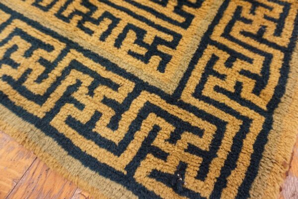 Golden yellow, high-pile rug with a dark blue interlocking key pattern rests on distressed wood planks.