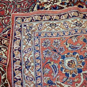 Corner of a low-pile rug folded back, detailing intricate red, navy, and cream floral borders.