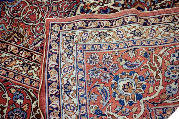 Corner of a low-pile rug folded back, detailing intricate red, navy, and cream floral borders.