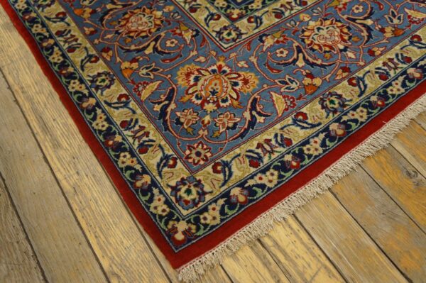 A low-pile, blue rug with detailed red and gold floral motifs lies on light, distressed wood planks.