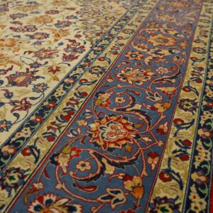 Ornate rug with an ivory field and wide blue border featuring red floral motifs on wood plank flooring.