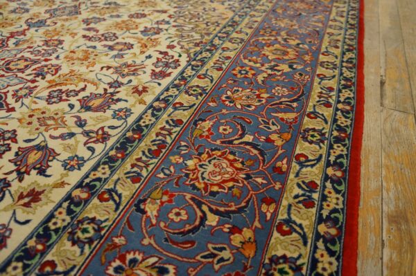 Ornate rug with an ivory field and wide blue border featuring red floral motifs on wood plank flooring.