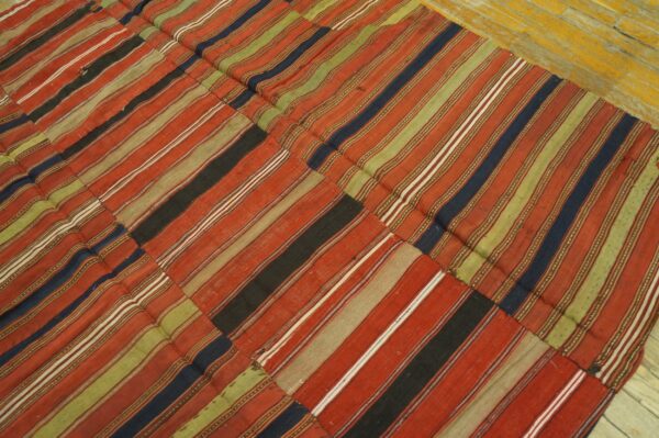 Patchwork low-pile rug featuring wide stripes in red, olive green, navy, and black on yellow wood flooring.