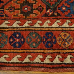 Red rug detail showing blue, teal, and gold geometric medallions bordered by cream wave designs on wood flooring.