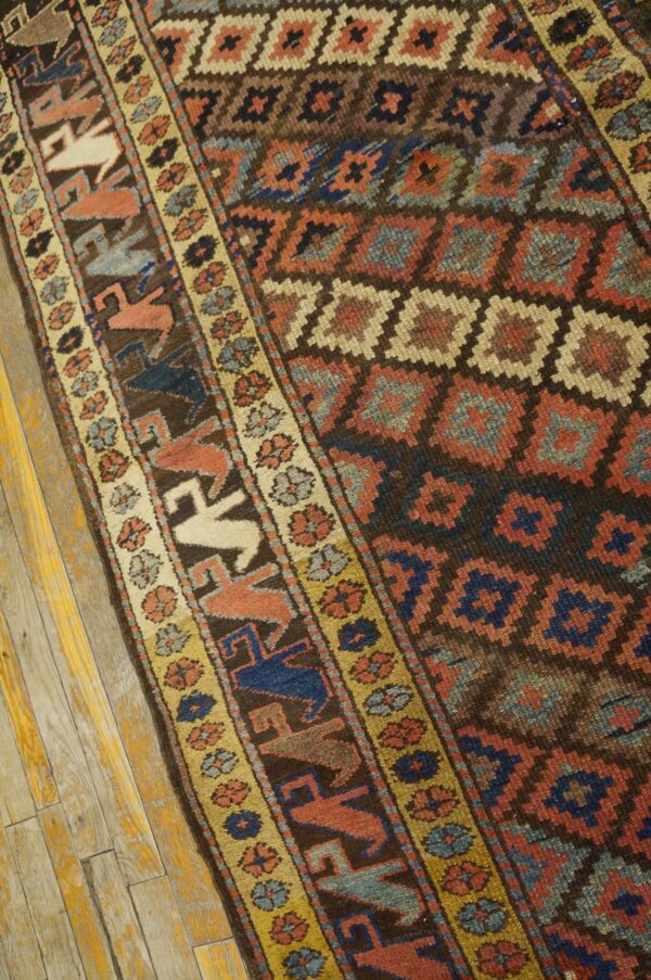 Rustic, medium-pile rug displaying dark diamond patterns, yellow floral borders, and stylized motifs on distressed wood flooring.