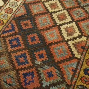Close-up shows a geometric diamond pattern in rust, blue, and beige against a dark brown field.