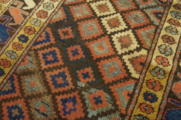 Close-up shows a geometric diamond pattern in rust, blue, and beige against a dark brown field.