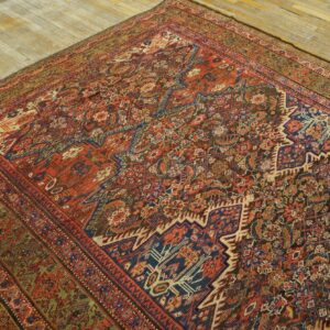Low-pile rug with intricate geometric and floral designs in deep red, blue, and brown tones on a worn wood floor.