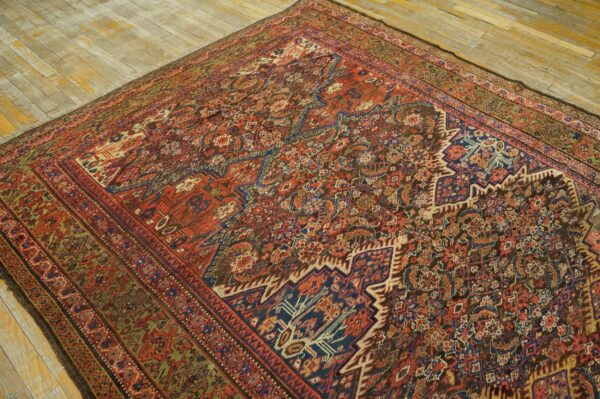 Low-pile rug with intricate geometric and floral designs in deep red, blue, and brown tones on a worn wood floor.