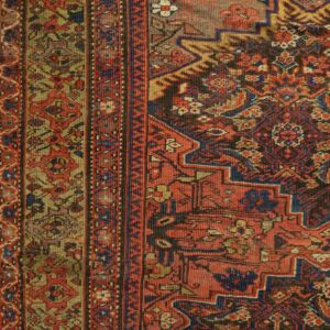Intricate traditional rug detail in red, blue, gold, and dark brown, resting on light wood planks.
