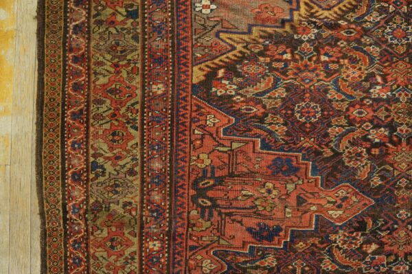 Intricate traditional rug detail in red, blue, gold, and dark brown, resting on light wood planks.