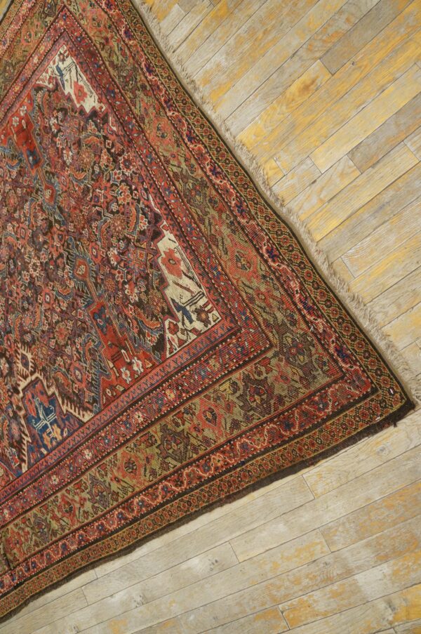 Muted red and olive traditional rug with intricate detail rests on distressed, light-colored wooden planks.