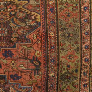 Worn, low-pile rug in red, blue, and khaki, displaying intricate geometric patterns on chipped yellow floorboards.
