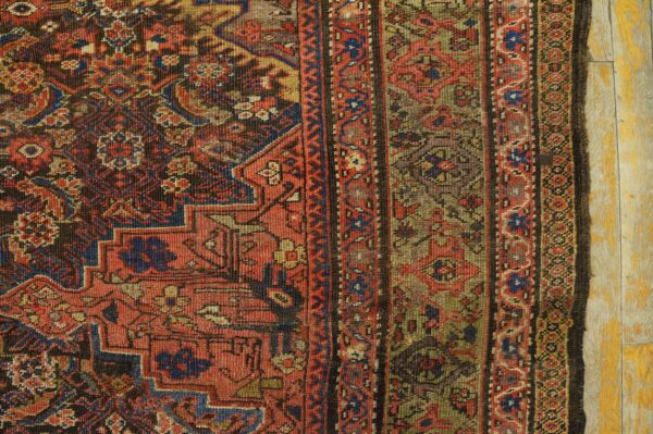 Worn, low-pile rug in red, blue, and khaki, displaying intricate geometric patterns on chipped yellow floorboards.