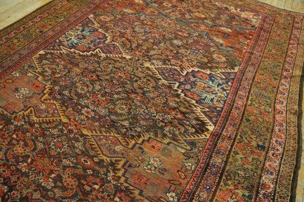 Detailed rust, blue, and green patterned rug with a wide olive border laid on pale wood flooring.