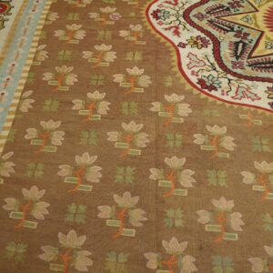 Detailed low-pile rug with a brown background, bright floral motifs, and elaborate borders in red, teal, and cream.
