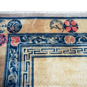 Tan and dark blue rug corner showing a geometric key border, scrolling vines, and round red floral motifs.