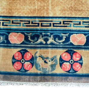 Tan rug detail with borders featuring navy and light blue Greek key, coral florals, and a central animal motif.
