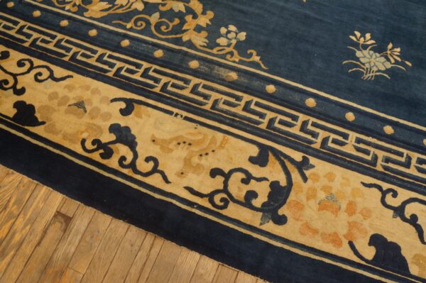 Navy blue rug with wide tan borders detailed with florals and a geometric fret pattern over wood floors.