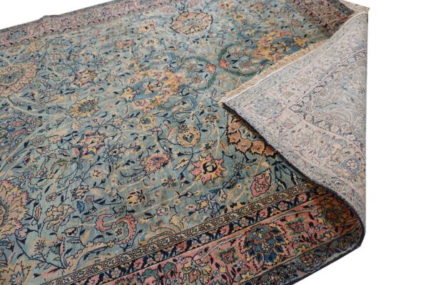 Ornate light blue rug with dense floral patterns and pink borders, partially folded to show the backing and fringe.