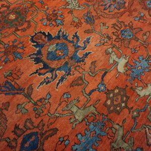 Low-pile rug detail showing an orange field with elaborate motifs in blue, olive, and brown.