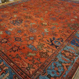 An orange-red rug with intricate floral motifs and a wide blue border lies on rustic wooden floors.