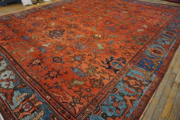An orange-red rug with intricate floral motifs and a wide blue border lies on rustic wooden floors.