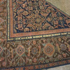 Close-up of a traditional rug corner showing dark brown, navy, coral, and blue patterns on light wood flooring.