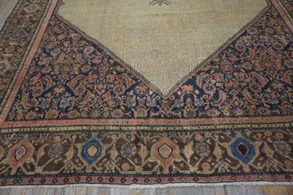 Corner of a traditional rug showing a beige geometric field meeting a border of navy, rust, and brown florals on wood planks.