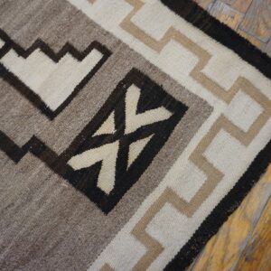 Flatwoven rug with stepped geometric patterns in cream, tan, gray, and dark brown on distressed wood flooring.