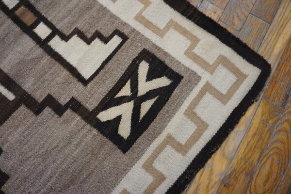 Flatwoven rug with stepped geometric patterns in cream, tan, gray, and dark brown on distressed wood flooring.