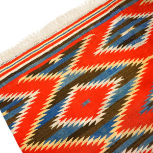 Flatwoven rug with a bold geometric pattern of red, blue, brown, and white zigzags, featuring white fringe.
