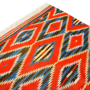 Close-up of a flatweave rug with red, blue, and brown serrated diamond patterns and white fringe.