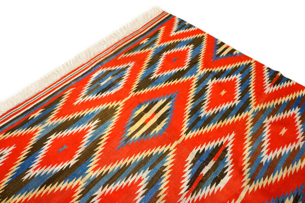 Close-up of a flatweave rug with red, blue, and brown serrated diamond patterns and white fringe.