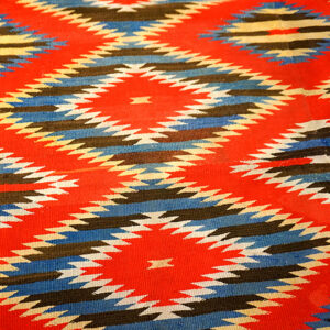 Flat-weave rug with geometric patterns of red, blue, black, and cream serrated diamonds.