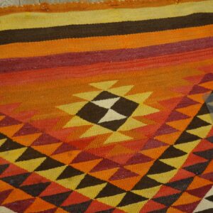 Geometric flat-weave rug featuring orange, red, yellow, and brown colors lying on worn wooden floors.